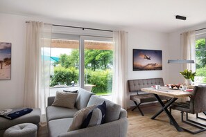 A modern living room with a sofa, a dining table with chairs, a bench, and a large window with a view of greenery.