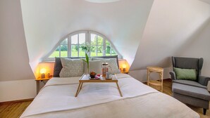 Une chambre avec un grand lit, une chaise, une petite table avec un plateau, et une fenêtre donnant sur un paysage verdoyant.