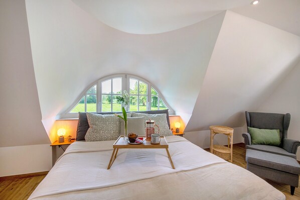 A bedroom with a large bed, a chair, a small table with a tray, and a window with a view of greenery.