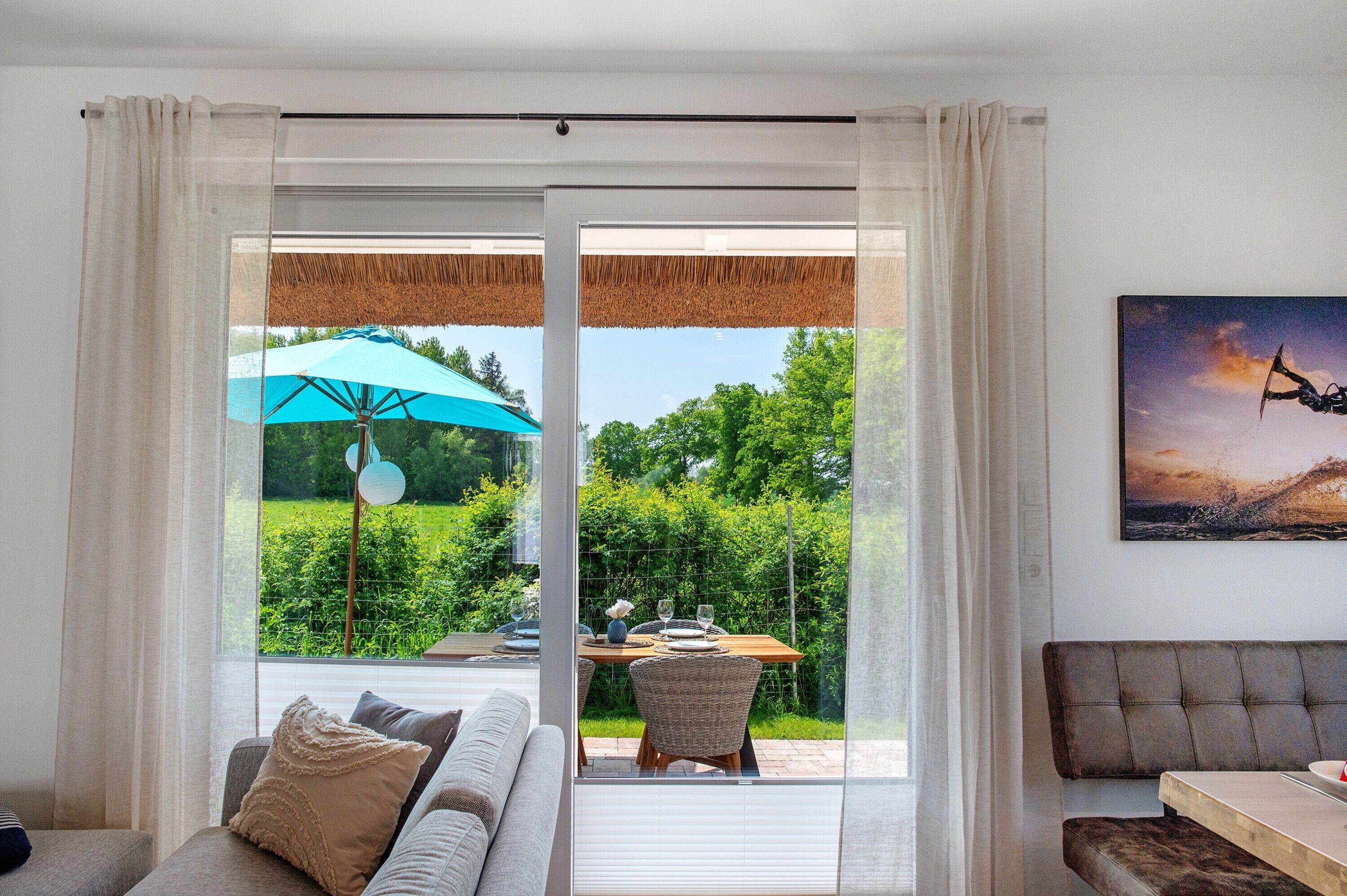 A living room with a sofa, a dining table with chairs, a large window with curtains, and a view of a garden.