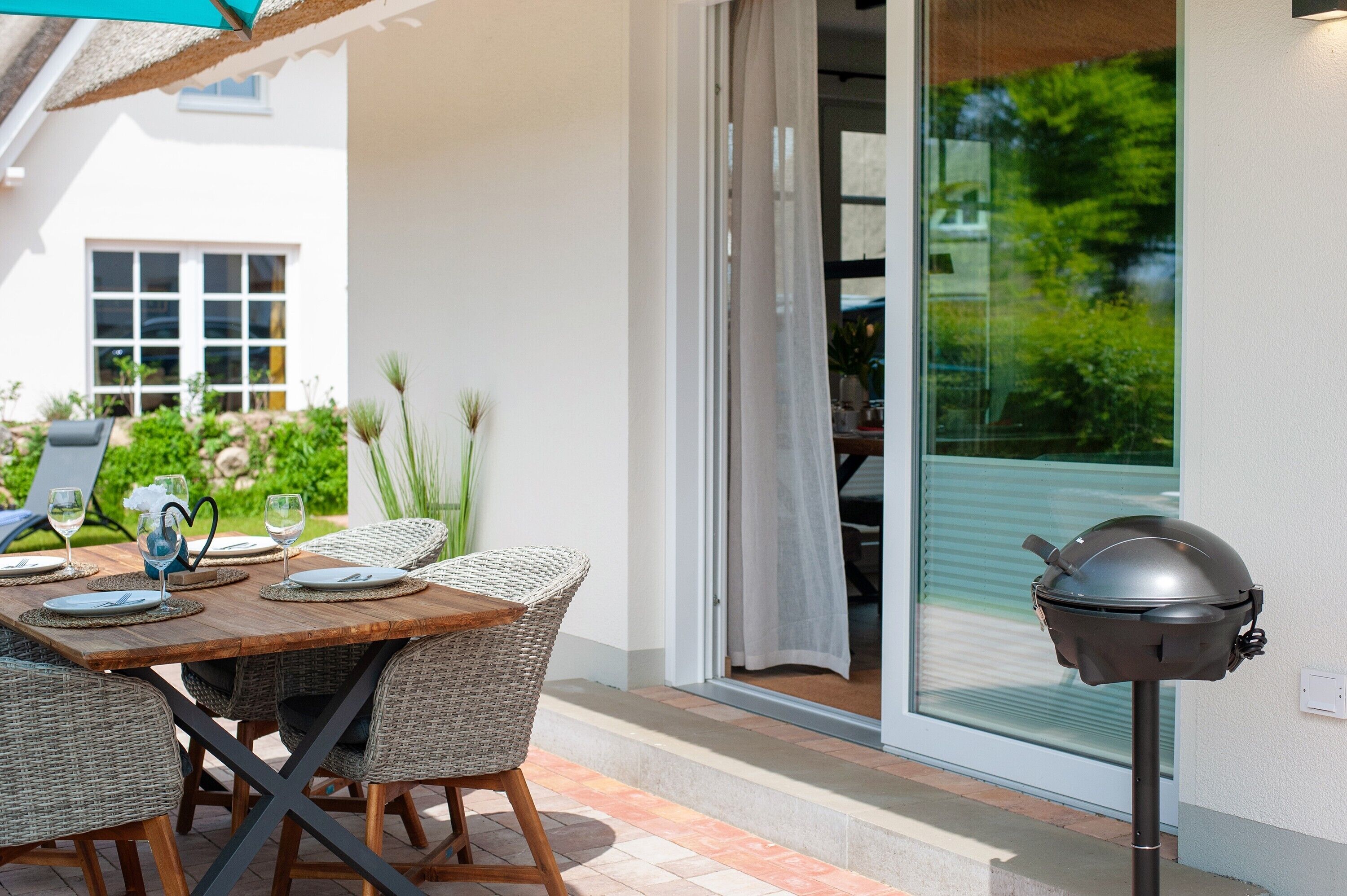 An outdoor dining area with wicker chairs and a wooden table, a grill, and a view of greenery through a sliding glass door.