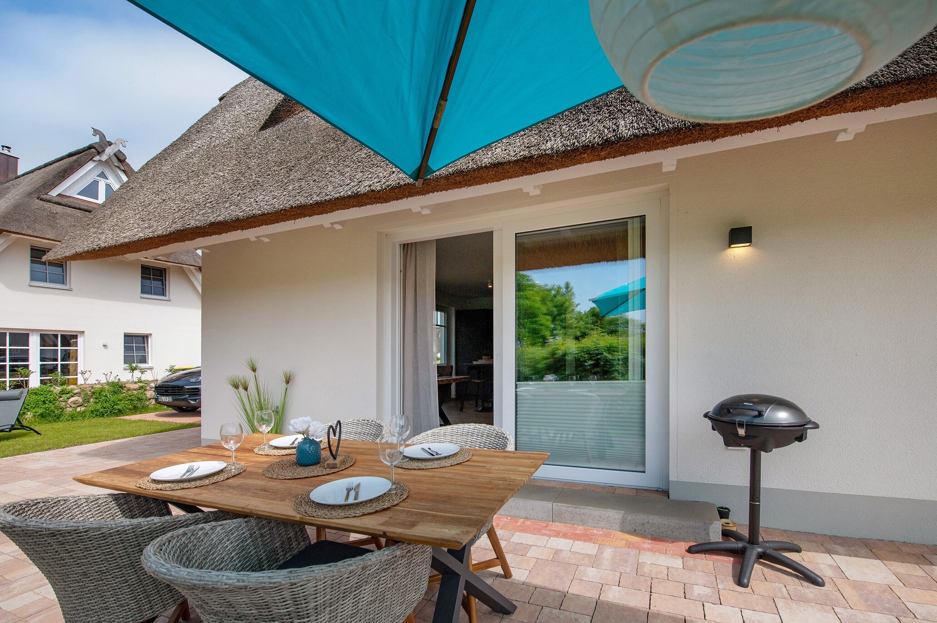 An outdoor patio with a wooden table set for dining, wicker chairs, a barbecue grill, and a blue canopy.