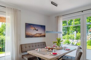 A dining area with a table set for two, a bench, a painting, and a view of greenery outside.
