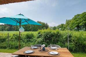 An outdoor dining table with a blue umbrella, surrounded by greenery and a wire fence.