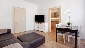 Uma sala de estar moderna com sofá, televisão em um móvel, mesa de jantar com cadeiras e cozinha visível por uma porta aberta.