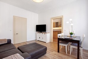 A modern living room with a sofa, a TV on a cabinet, a dining table with chairs, and a kitchen visible through an open door.