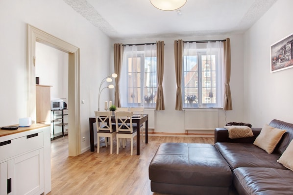 Un salon moderne avec un canapé en cuir marron, une table à manger avec des chaises, un coin cuisine équipé d’un four  à micro-ondes et de grandes fenêtres avec des rideaux.