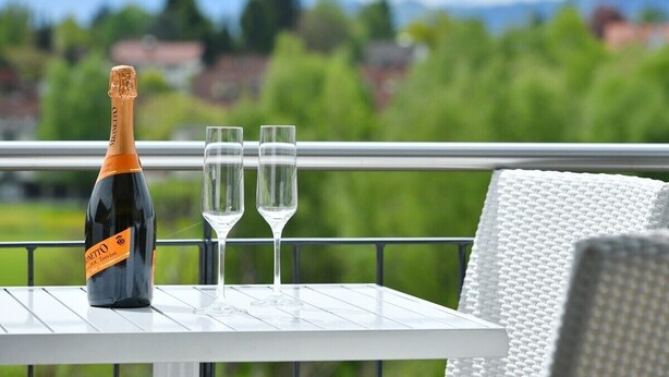 Una botella de vino, dos copas y una mesa blanca en un balcón con vistas a zonas verdes y casas.