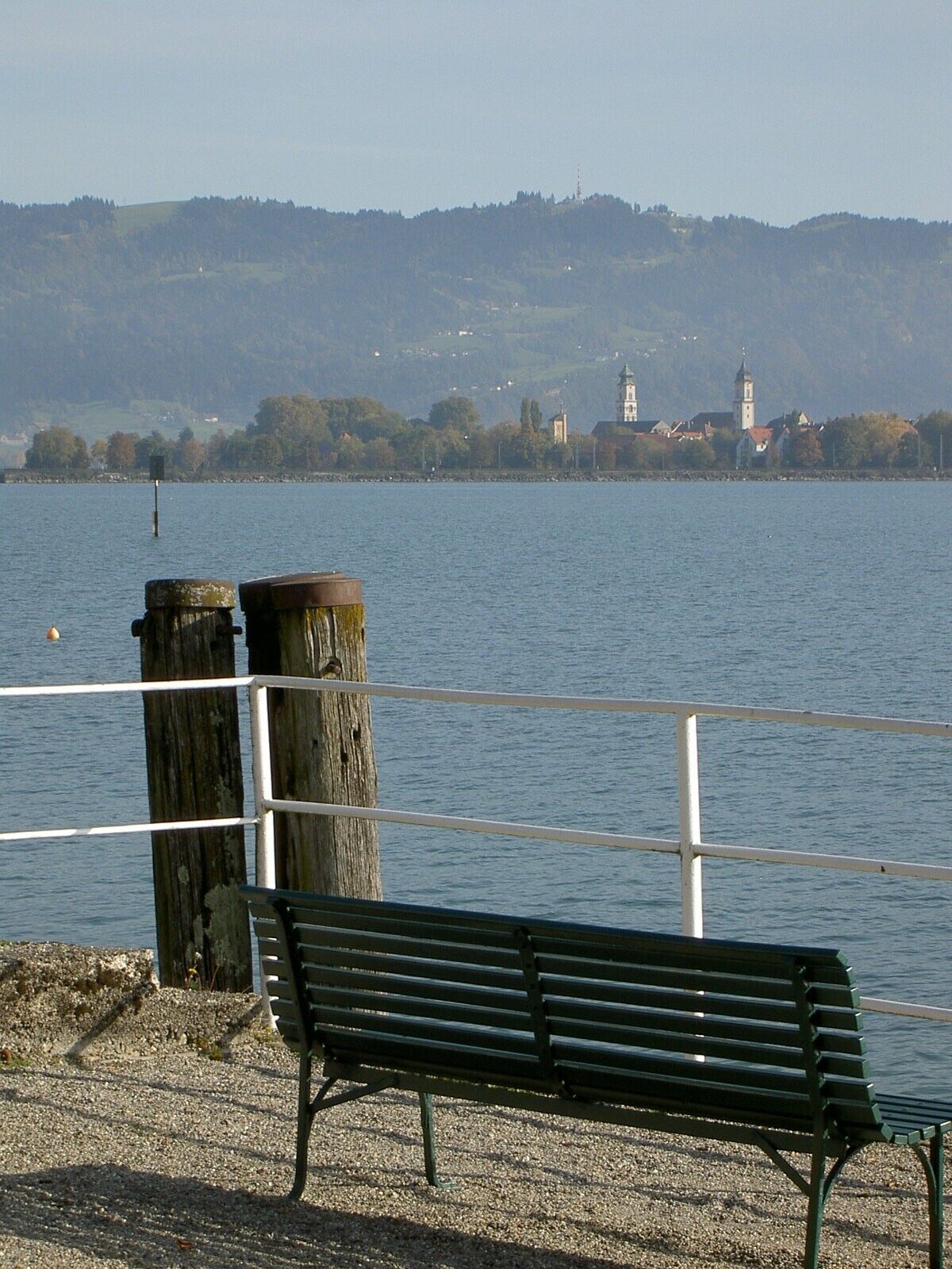 Eine Bank am Wasser mit Blick auf eine Stadt und Hügel in der Ferne.