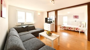 A living room with a grey sofa, a white coffee table with a fruit bowl, a TV on a white cabinet, and a bedroom with a floral bedspread. - Holiday Apartment 'Dinkelbach Landhaus 1 A Charmant' with Lake View, Balcony and Wi-Fi (Bodolz)
