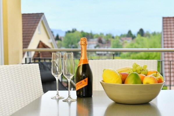 A table with a bottle of champagne, two glasses, and a bowl of fruit on a balcony with a view of houses.