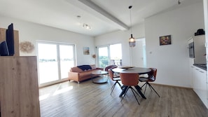 A spacious living area with a sofa, round dining table, and chairs, large windows, and a kitchen area.
