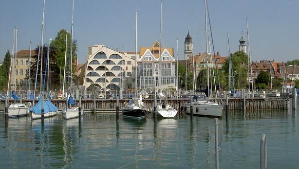 A marina with multiple boats docked, a distinctive building with a unique facade, and a clear sky.