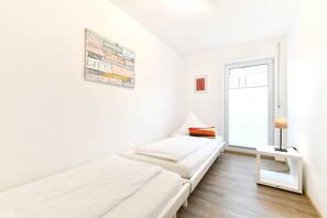 A small, bright room with two single beds, a wooden floor, a white wall with a word art piece, a small table with a lamp, and a door leading outside.