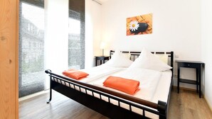 A double bed with white linens and orange throw pillows, a nightstand, a wall-mounted artwork, and a large window with a view of buildings.
