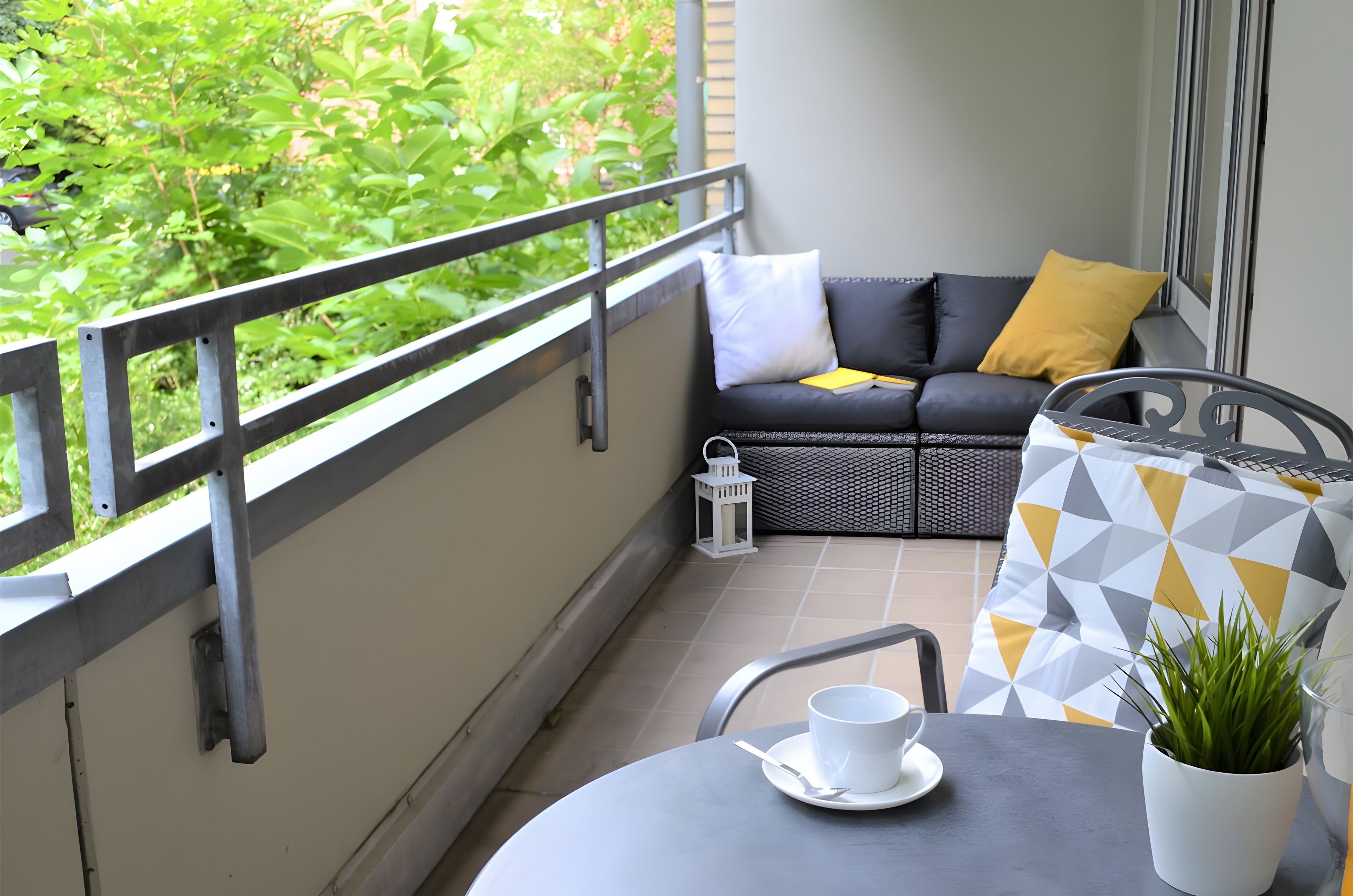En balkon med en sort sofa, gul pude, hvide hynder, en hvid lanterne og en potteplante.
