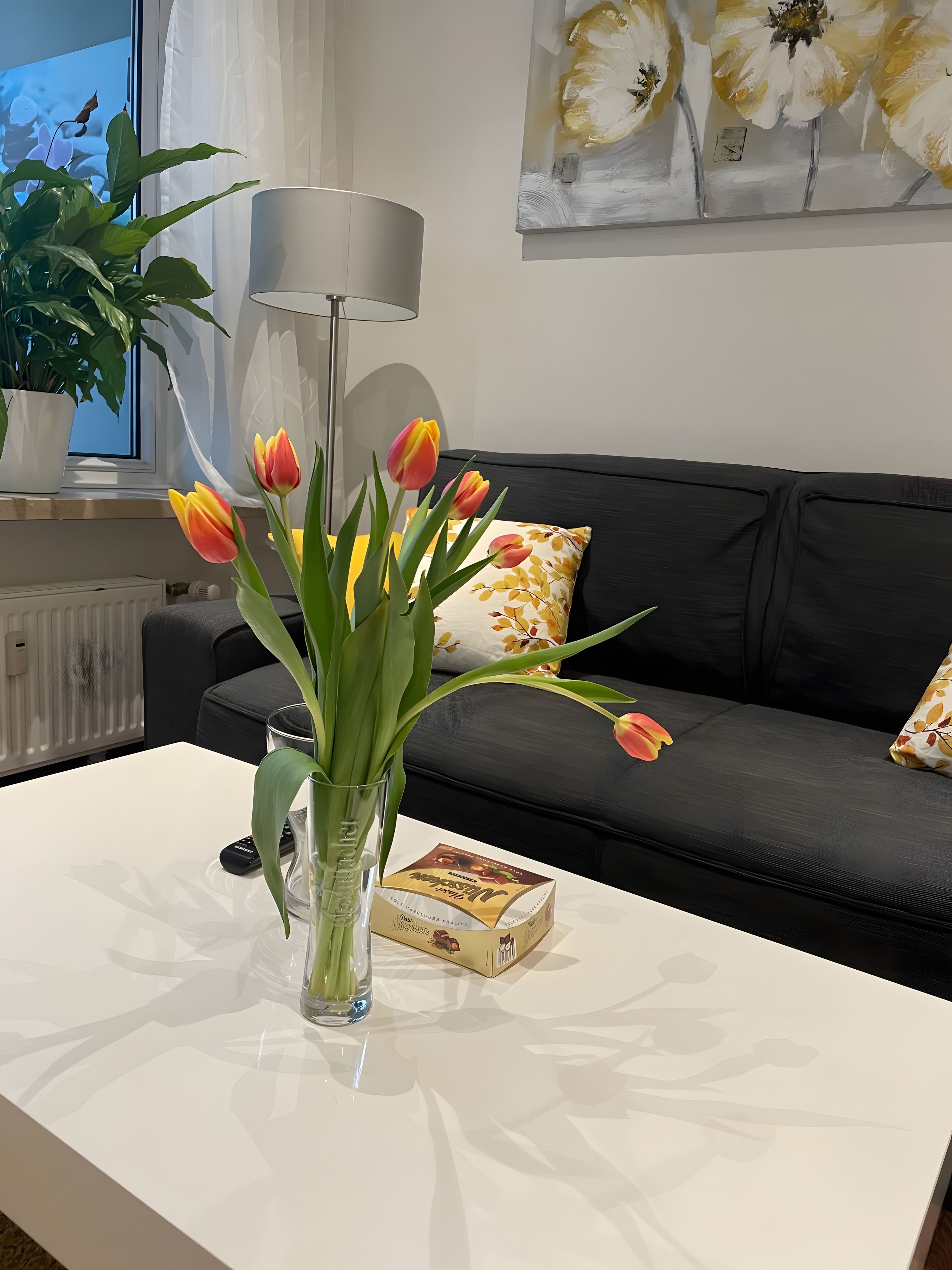 A modern living room with a white coffee table, a vase with tulips, a remote control, and a box of chocolates.