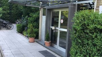 Um edifício com entrada de vidro, plantas em vasos e estrutura metálica sobre a porta.