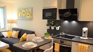 A modern kitchen with a microwave, oven, and coffee maker, a dining area with a table and chairs, and a living room with a sofa and cushions.