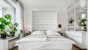 A modern bedroom with a large bed, bedside tables, a built-in shelving unit, and potted plants.