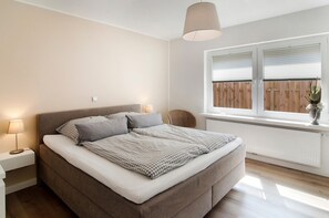 Une chambre à coucher avec un lit, des lampes de chevet, une fenêtre avec des stores, un radiateur et un fauteuil en osier.