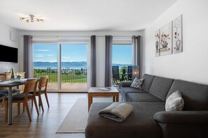 Una sala de estar moderna con un sofá, una mesa de comedor con sillas y un televisor. Hay un ventanal con vistas a un paisaje lejano.