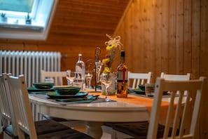 Une table à manger est dressée avec des assiettes, des verres et des bouteilles, entourée de chaises blanches dans une pièce aux murs en bois et au plafond en pente.