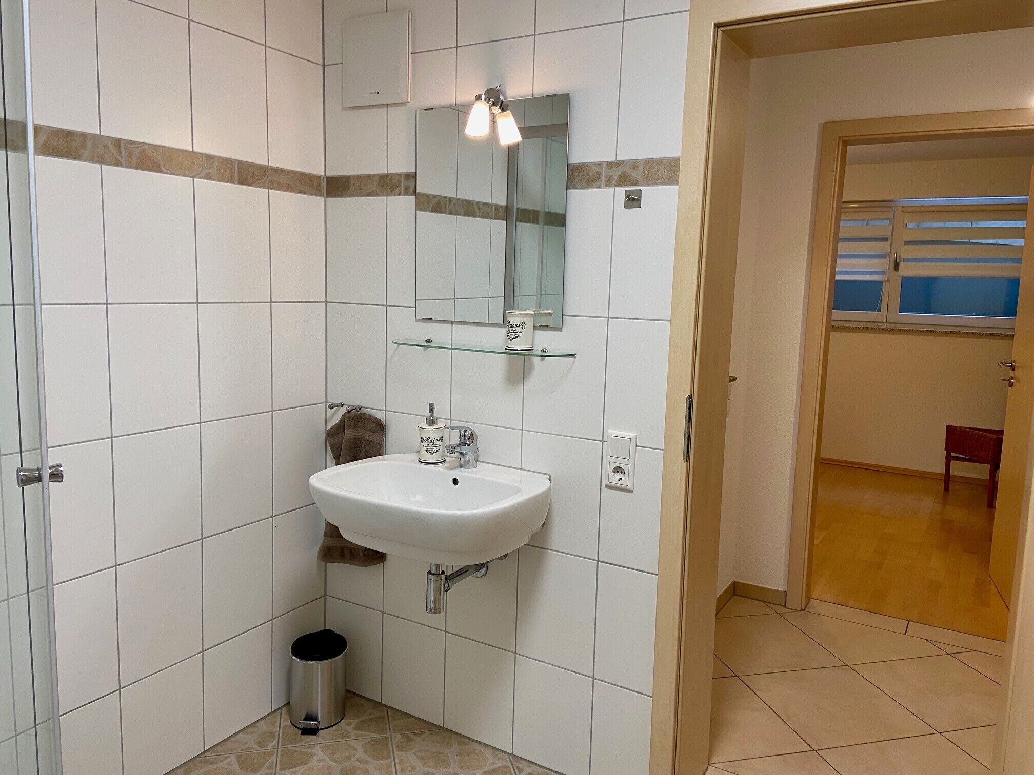 A bathroom with white tiled walls, a white sink, a glass shelf, a mirror, and a trash can.