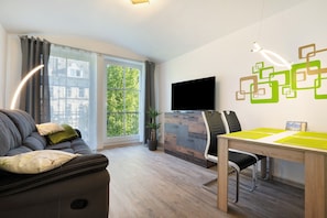 A modern living room with a sofa, a dining table, a flat-screen TV, and a large window with a view of greenery.
