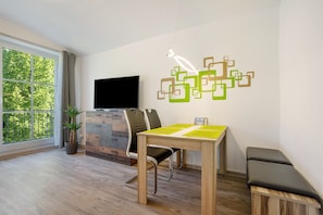 A modern living room with a TV, a dining table, chairs, a bench, a potted plant, and a large window.