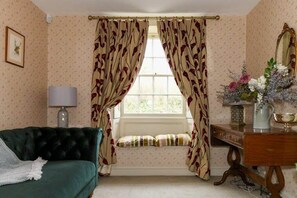 A room with a green sofa, a window with floral curtains, a lamp, and a dresser with a mirror and decorative items. - Beautiful Georgian Cottage near Frome (Beckington)