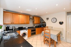 A kitchen with wooden cabinets, a washing machine, a dining table with chairs, and a clock on the wall. - Beautiful Georgian Cottage near Frome (Beckington)