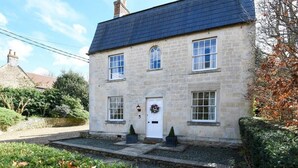A two-story stone house with a white door, multiple windows, and a small garden area with shrubs and a pathway. - Beautiful Georgian Cottage near Frome (Beckington)