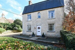 Une maison en pierre à deux étages, avec un toit sombre, une porte d’entrée blanche et un petit porche agrémenté de deux plantes en pot.
