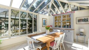 A conservatory with a wooden table and chairs, large windows, and a cabinet with glass doors. - Beautiful Georgian Cottage near Frome (Beckington)