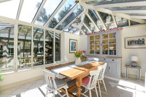 A conservatory with a wooden table and chairs, large windows, and a cabinet with glass doors.