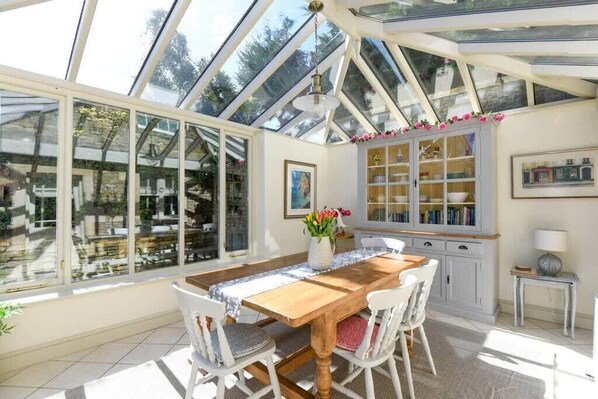 A conservatory with a wooden table and chairs, large windows, and a cabinet with glass doors. - Beautiful Georgian Cottage near Frome (Beckington)