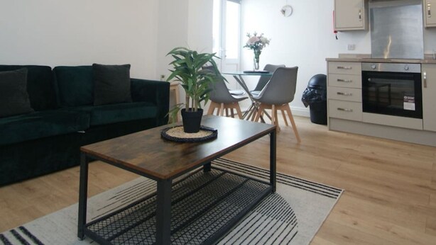 A modern living room with a dark green sofa, a wooden coffee table, and a potted plant. There is a dining area with a glass table and chairs, and a kitchen with white cabinets and stainless steel appliances.