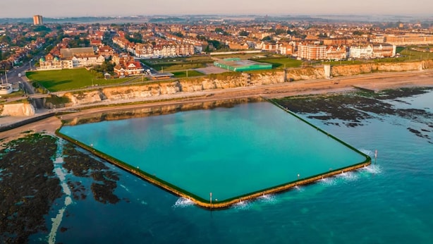 Vue aérienne d’une zone côtière avec un grand plan d’eau rectangulaire, une promenade et des immeubles résidentiels en arrière-plan.