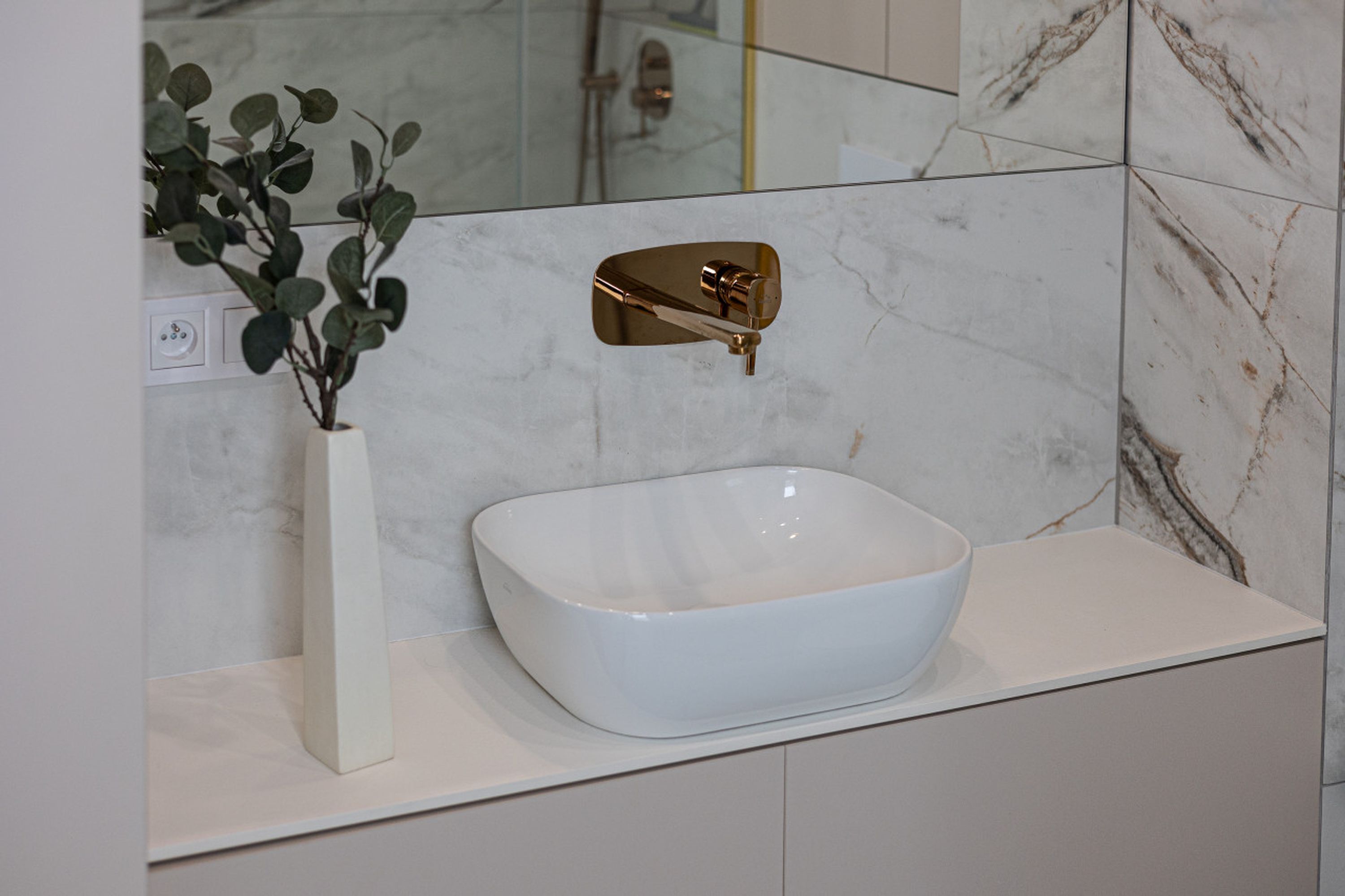 Une salle de bain avec un lavabo en marbre blanc, un robinet doré et un vase blanc contenant des feuilles vertes.