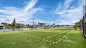 Cancha de tenis al aire libre con red, rodeada por una cerca, con vista a un muelle con edificios y una grúa al fondo.