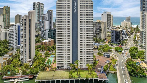 Un edificio de gran altura con un letrero llamativo que dice 'CONDOR', rodeado de otros edificios altos, un puerto deportivo y una carretera con vehículos.