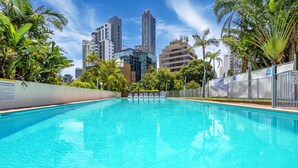 Una piscina con agua azul cristalina, rodeada de palmeras y un paisaje urbano al fondo.