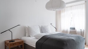 A minimalist bedroom with a bed, bedside tables, a lamp, and a window with sheer curtains. - Queens Courtyard 2BR XL (København)