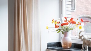 A room with a window, a curtain, a vase with flowers, and a small elephant figurine on the windowsill. - Queens Courtyard 2BR XL (København)