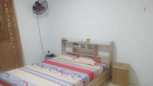 Un dormitorio con cama, mesita de noche, estante pequeño, ventilador y un cesto de basura.