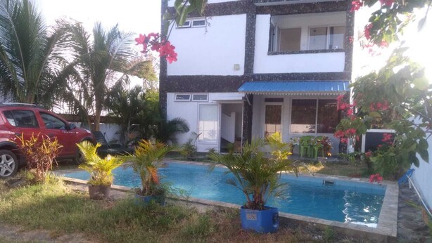 Un edificio de dos pisos con piscina en el jardín delantero, rodeado de palmeras y plantas en macetas.