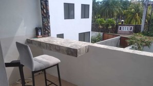 Un balcón con un taburete, una mesita y vistas a edificios y zonas verdes cercanas.