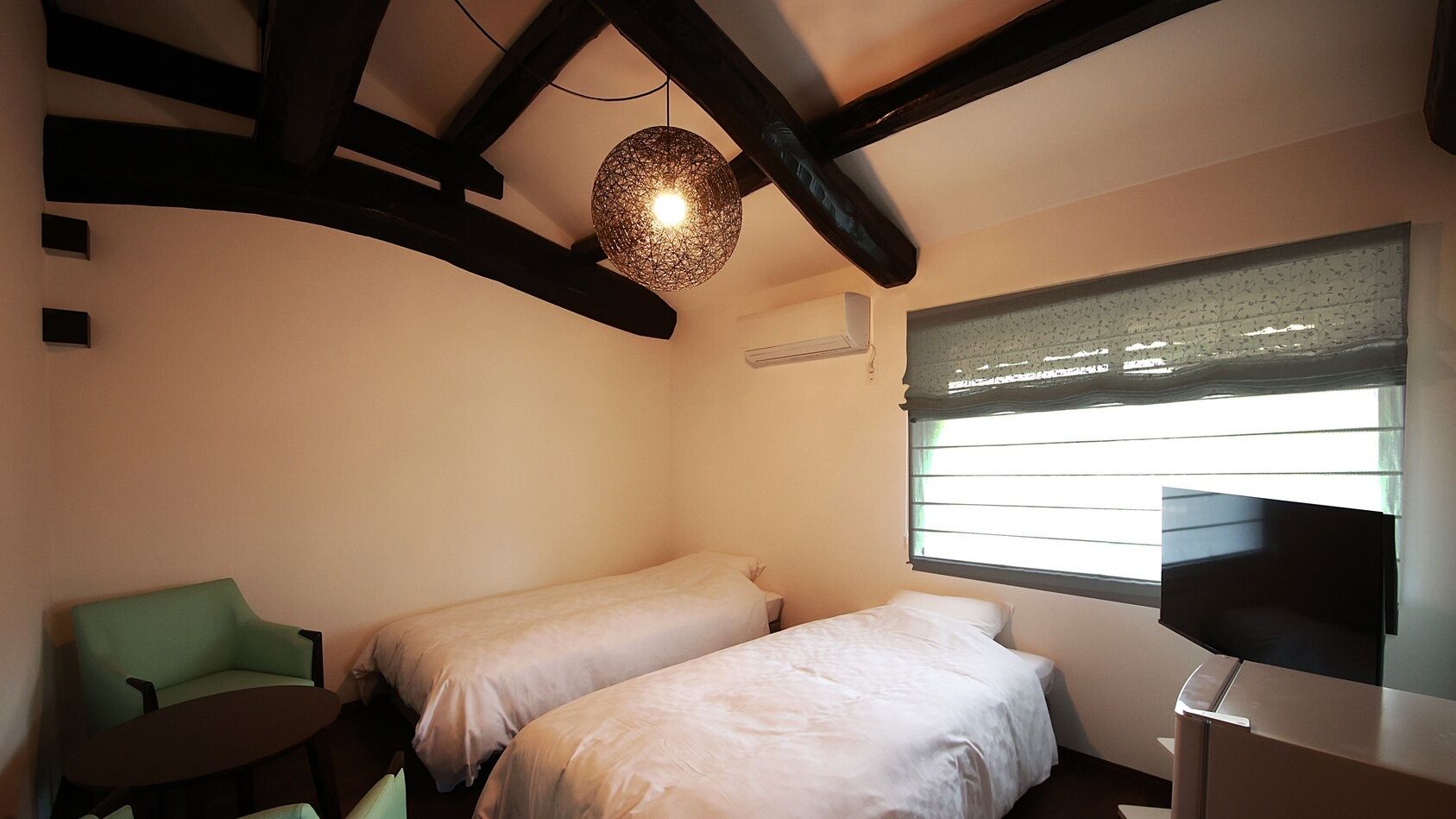 A bedroom with two beds, a green chair, a table, a TV, and a hanging light fixture.