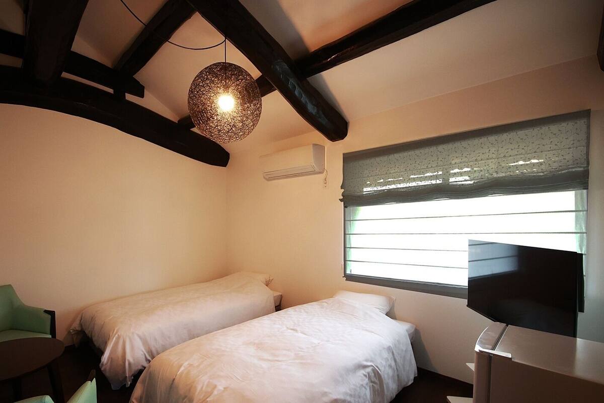Un dormitorio con dos camas, una silla verde, una mesa, un televisor y una lámpara colgante.